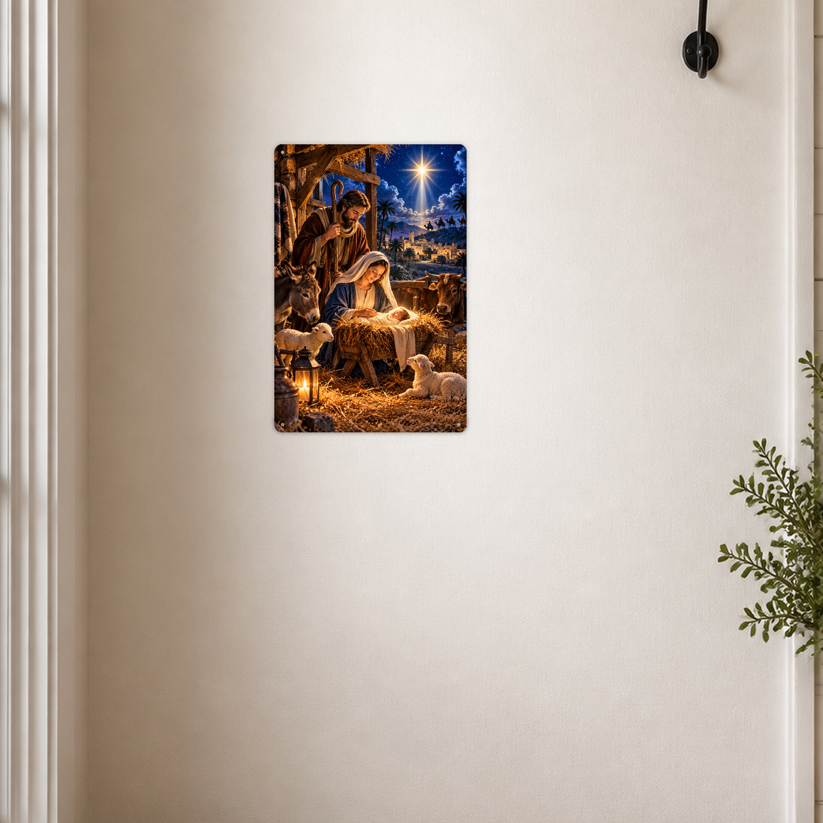 The aluminum sign showing the Holy Family at the manger with a donkey, ox, and lambs, mounted on a cream wall near a coat hook and a potted plant.