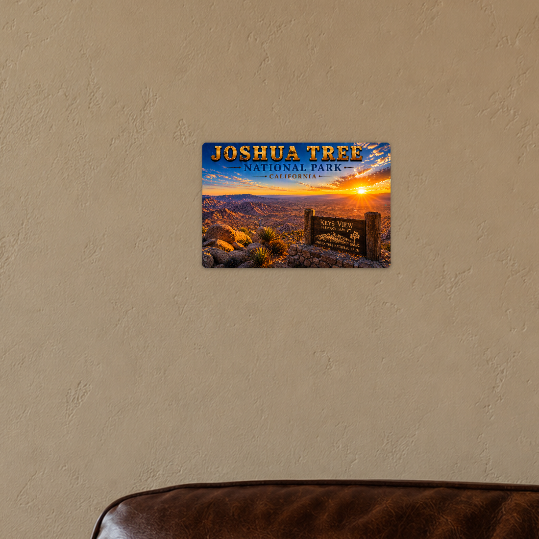 Sign showing Joshua Tree Keys View lettering and a sunset desert lookout, displayed on a warm tan plaster wall above the corner of a brown leather chair.