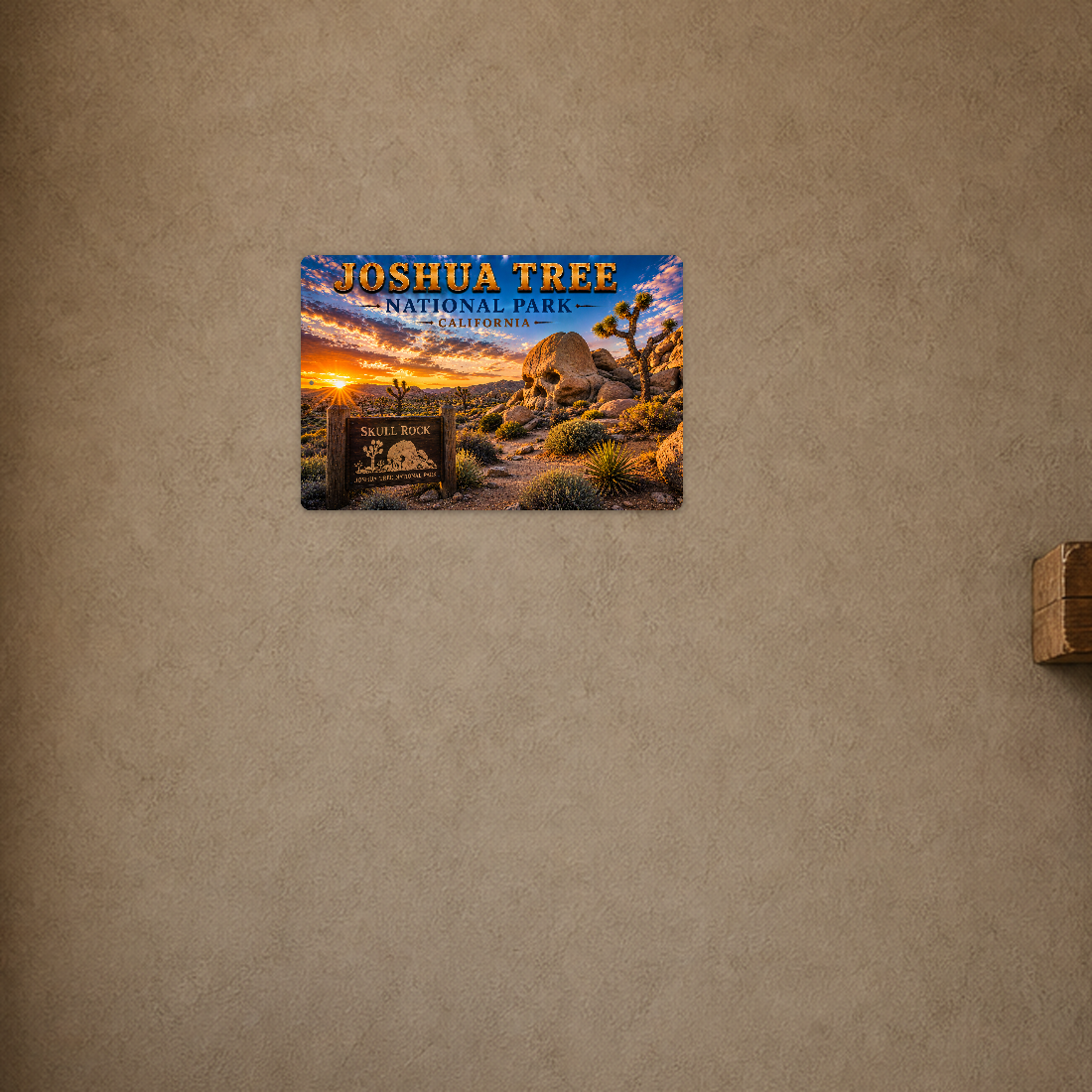 Sign showing Joshua Tree Skull Rock lettering with boulders and Joshua trees at sunset, displayed on a warm tan wall with part of a wooden cabinet below.
