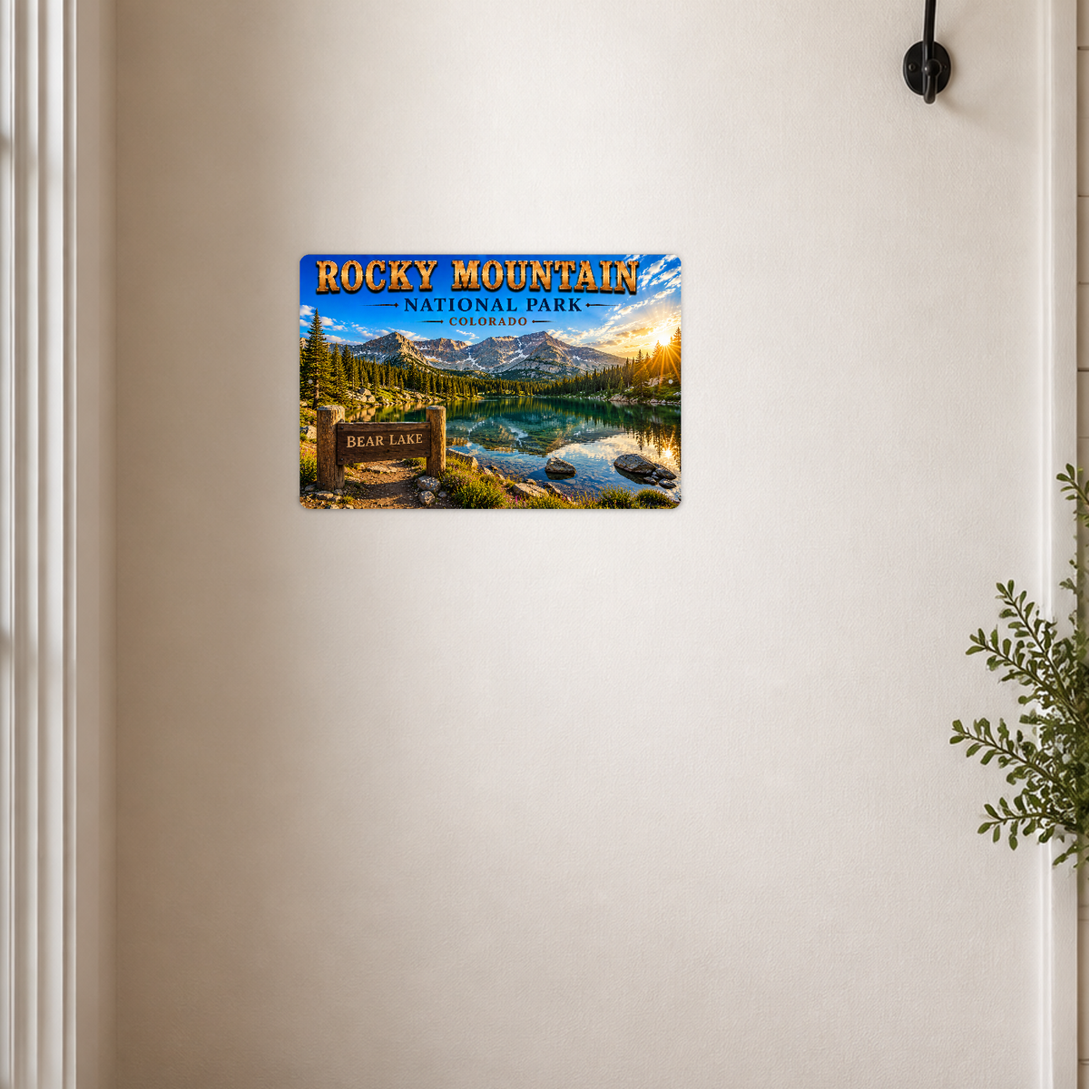 Sign showing Rocky Mountain Bear Lake lettering and a turquoise lake below snowy mountains, displayed on a cream wall beside a white-framed doorway and black wall fixture.