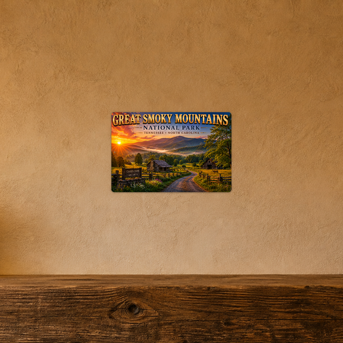 Sign showing Great Smoky Mountains Cades Cove lettering with cabins and a fenced valley road at sunset, displayed on a warm tan textured wall above a wooden ledge.