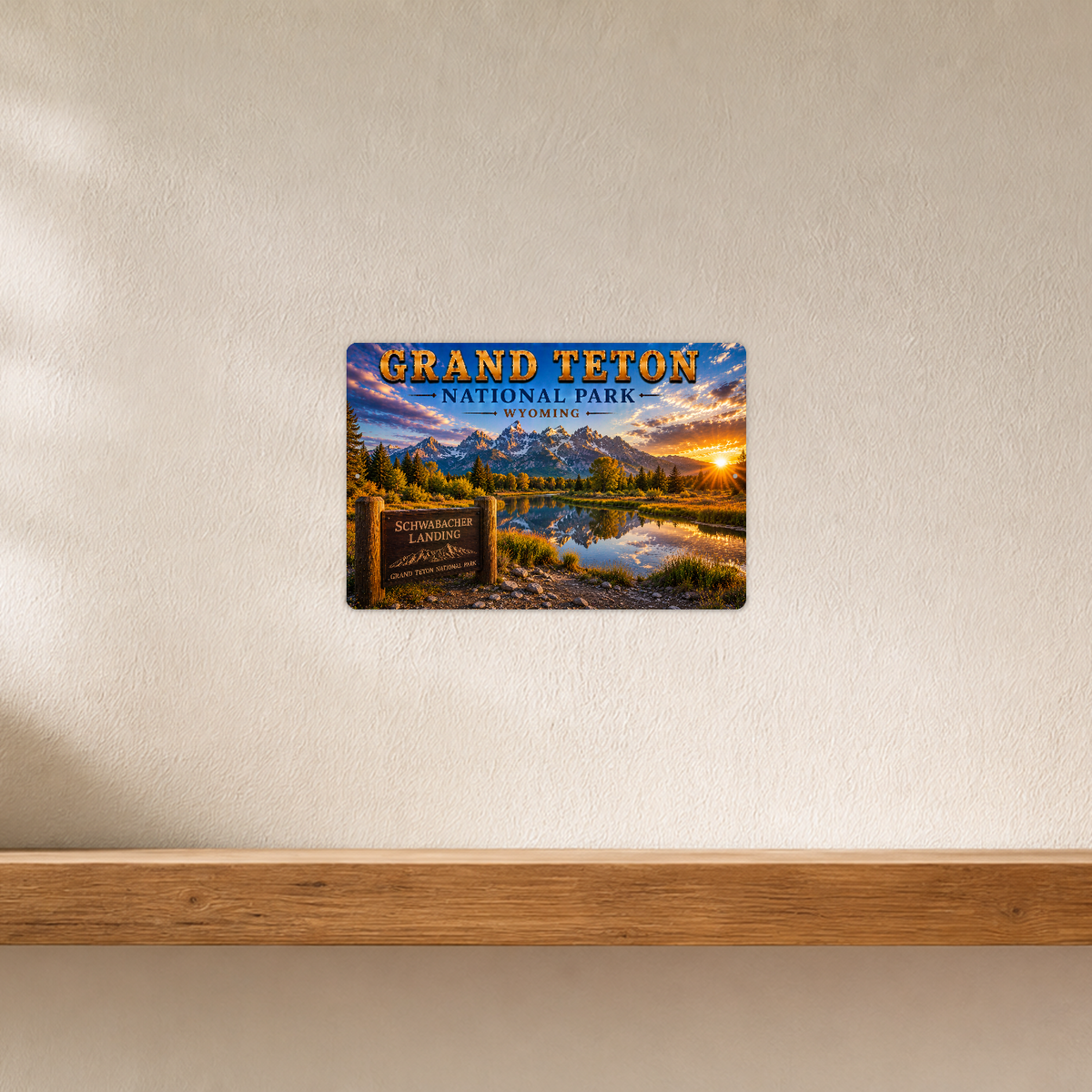 Sign showing Grand Teton Schwabacher Landing lettering and mountains reflected in a sunset river, displayed on a white plaster wall above a horizontal wooden ledge.
