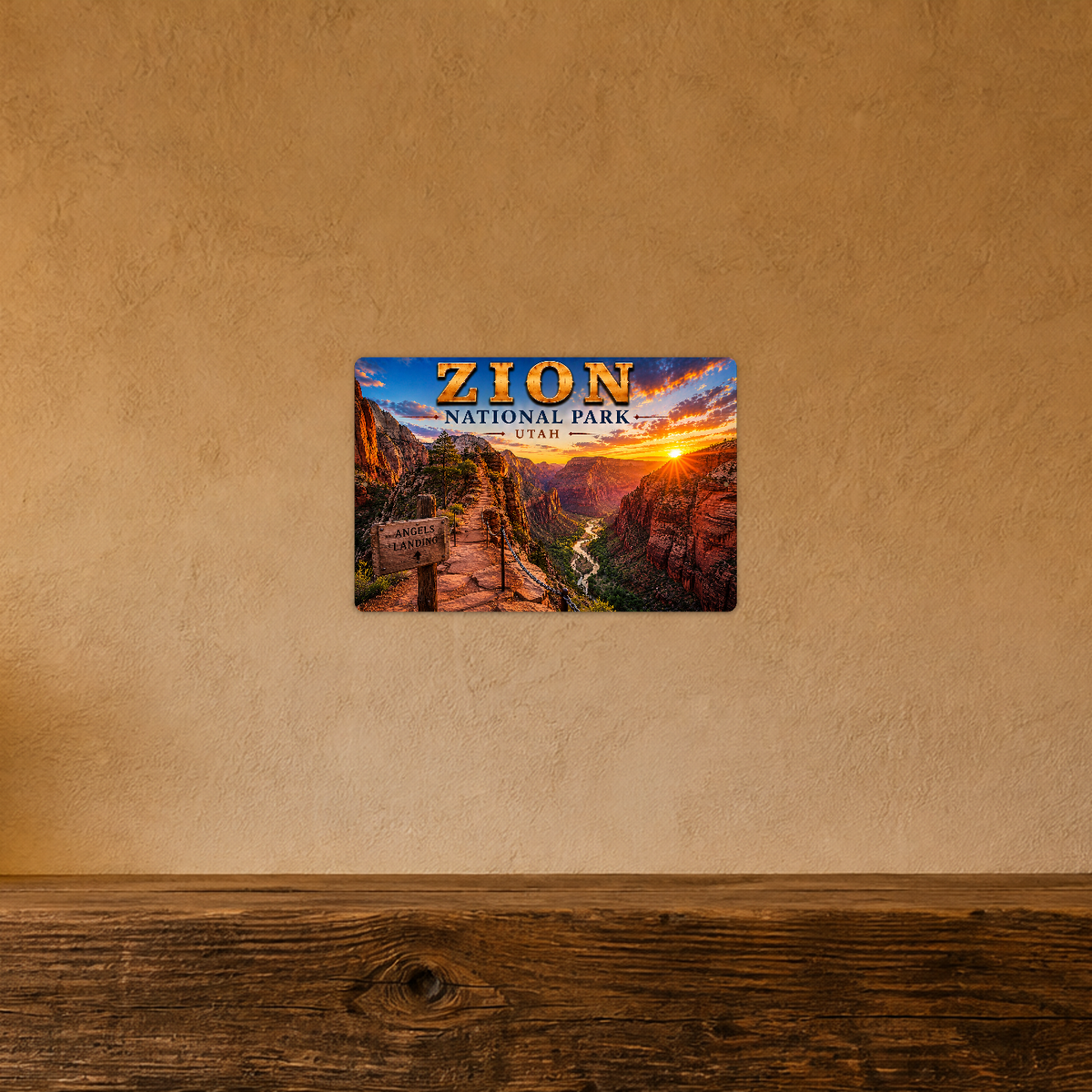 Sign showing Zion Angels Landing lettering with a path above a red canyon and river, displayed on a warm tan textured wall above a wooden ledge.