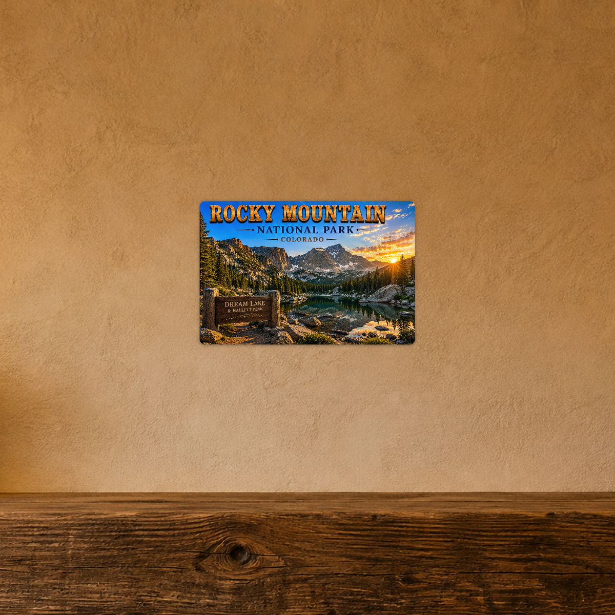 Sign showing Rocky Mountain Dream Lake Hallett Peak lettering with a mountain lake and pine forest, displayed on a warm tan textured wall above a wooden ledge.