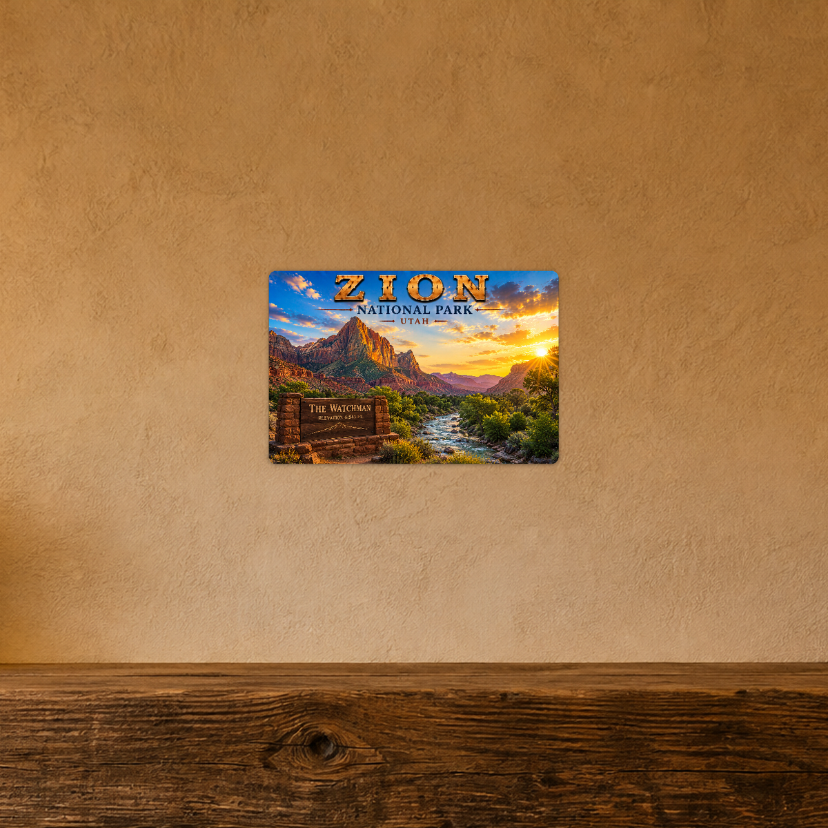 Sign showing Zion The Watchman lettering with a river, red mountain and forest, displayed on a warm tan textured wall above a wooden ledge.