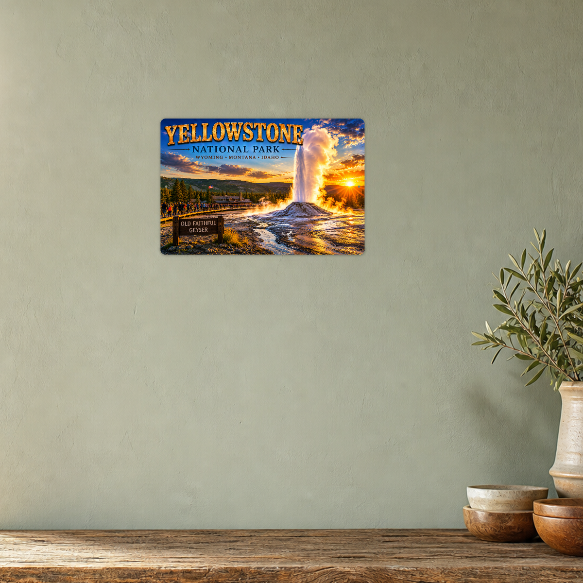Yellowstone Old Faithful sign showing an erupting geyser at sunset, displayed on a pale sage wall above a rustic wooden table with stacked bowls and a leafy plant in a tan ceramic vase.