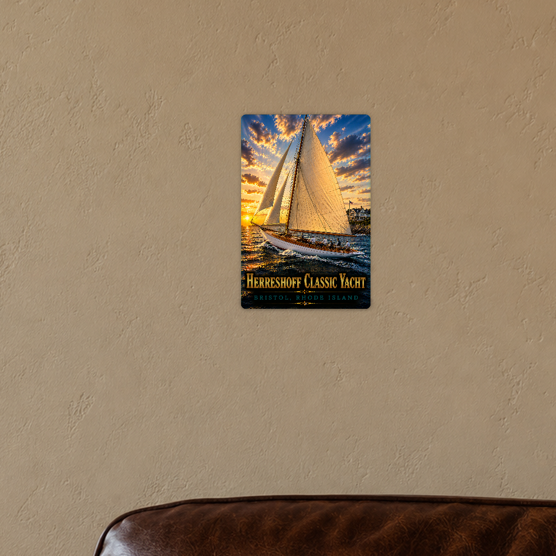 Sign showing Herreshoff Classic Yacht Bristol lettering and people aboard a white-hulled boat with golden sails, displayed on a warm tan wall above a brown leather chair corner.