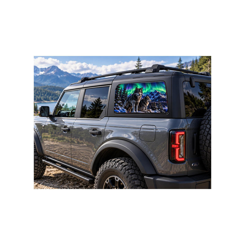 Ford Bronco rear side window with a perforated decal of two wolves standing on a snowy rock under green and purple northern lights, snow-capped mountains and pine trees in the scene, applied to the outside of the glass.