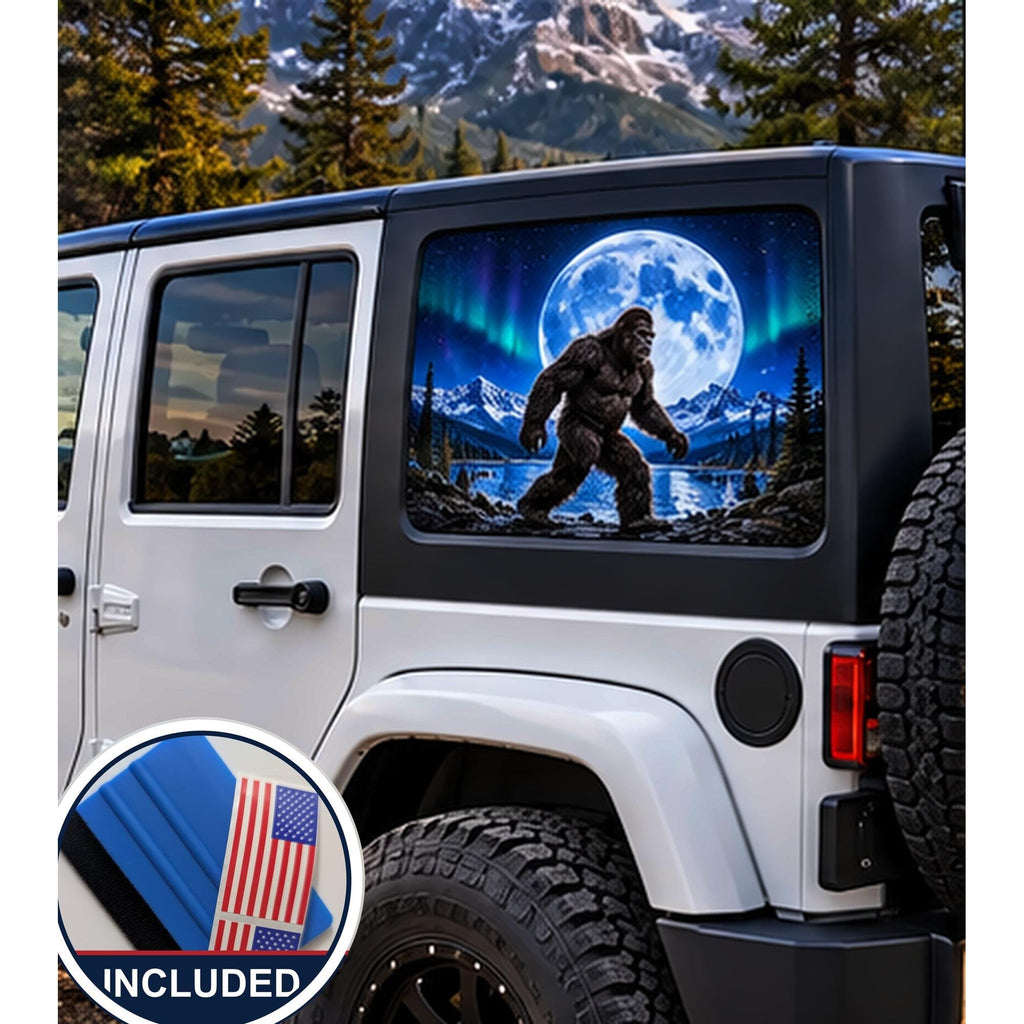 Jeep Wrangler 4-door rear quarter window with a perforated decal of Bigfoot walking beside a mountain lake under a full moon and green and purple northern lights, applied to the outside of the glass. An INCLUDED badge in the corner shows the felt squeegee and 2 American flag decals that come with every set.