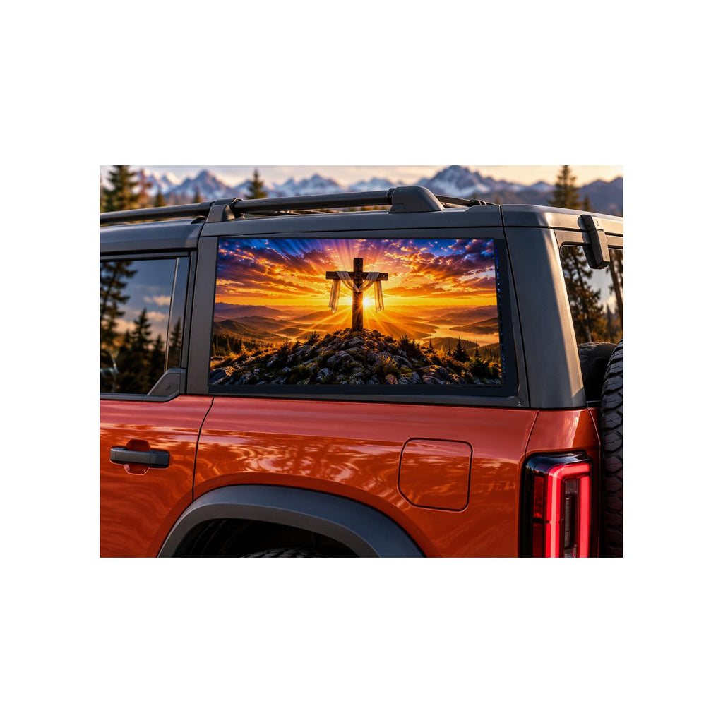 Ford Bronco rear side window with a perforated decal of a wooden cross draped with cloth on a mountaintop at sunrise, sun rays breaking through the clouds, applied to the outside of the glass.