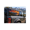 Ford Bronco rear side window with a perforated decal of a waving American flag over snow-capped mountains and a small pond at a fiery orange sunset, applied to the outside of the glass.