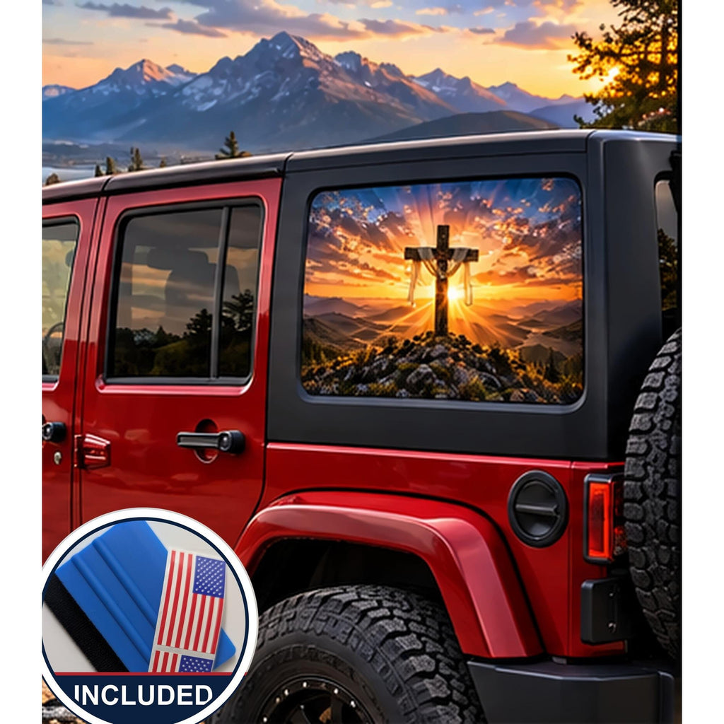 Jeep Wrangler 4-door rear quarter window with a perforated decal of a wooden cross draped with cloth on a mountaintop at sunrise, sun rays breaking through the clouds, applied to the outside of the glass. An INCLUDED badge in the corner shows the felt squeegee and 2 American flag decals that come with every set.